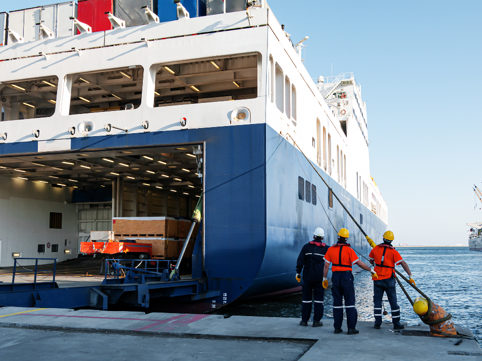 Container ship moored to the dock. The back of the ship is open, there are three crew wearing hard hats next to the ship.