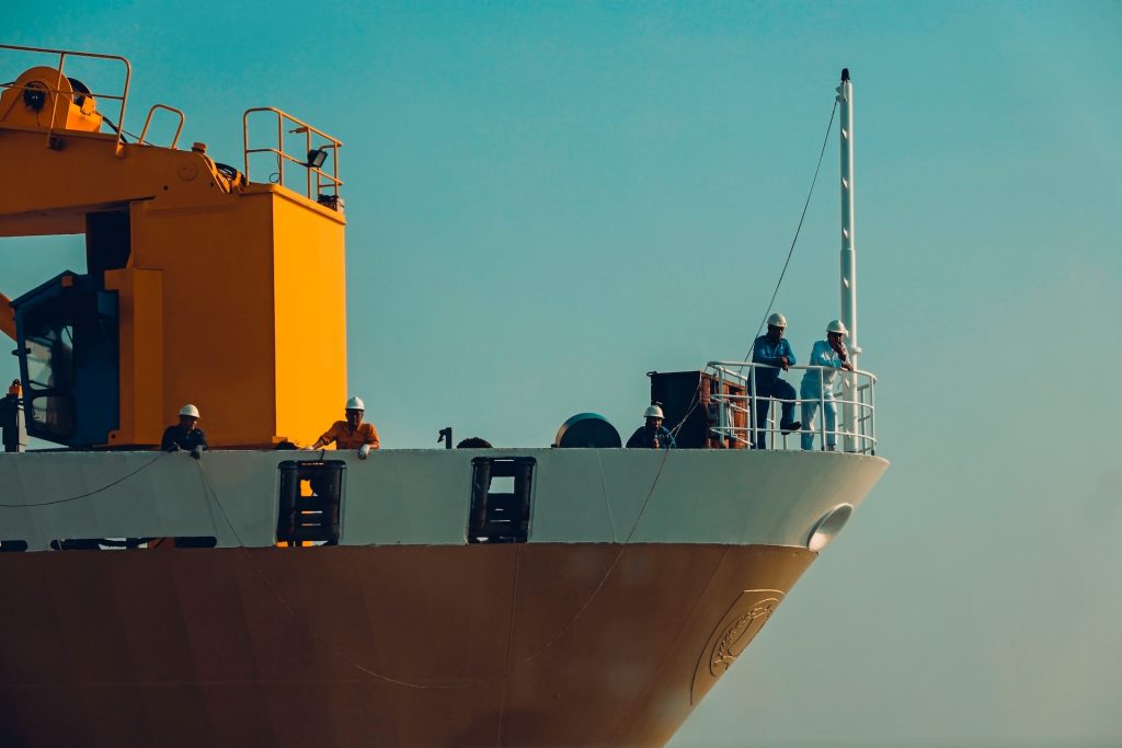 Bow of a container ship. It is yellow and white, with crew members visible looking over the side of the ship